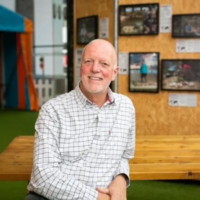A person smiling with very short white hair, they are wearing a white chequered shirt, and are sitting at a table in front of a We The Curious exhibit.