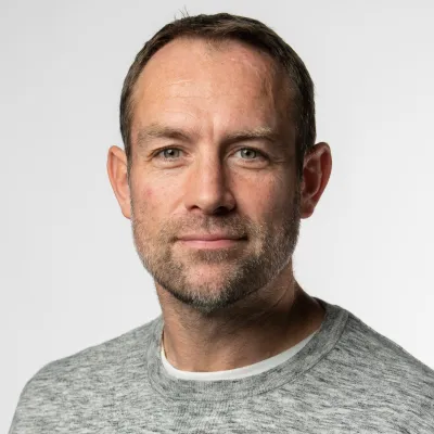 person smiling with short brown hair, they are wearing a grey jumper, and are in front of a plain background.
