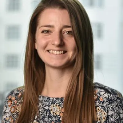 A person smiling with long brown hair, they are wearing a patterned top, and are in front of a plain background.