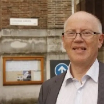 A person smiling with glasses, they are wearing a shift and grey suit jacket, and are in front of a building.