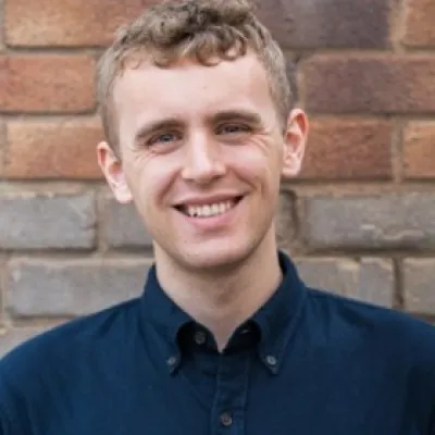 A person smiling with short blonde hair, they are wearing a navy shirt, and are in front of a brick background