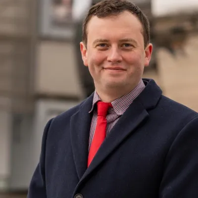 A person smiling with short brown hair, they are wearing a black suit jacket and a red tie, and are in front of a building.