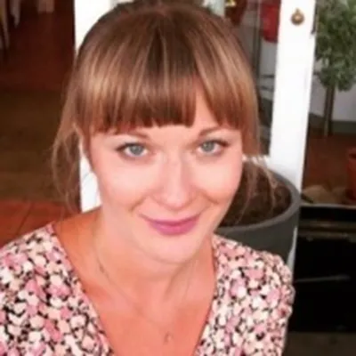 A person smiling with light brown hair tied back and a fringe. They are wearing a flowery print top and they are in front of a doorway.