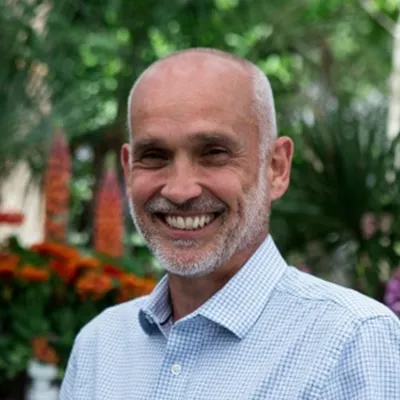 A person smiling with very short grey hair. They are wearing a blue shirt and are in front of trees.