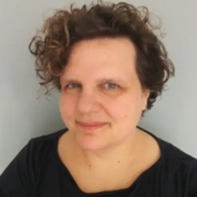 A person smiling with short curly brown hair. They are in front of a grey background and they are wearing a black top.