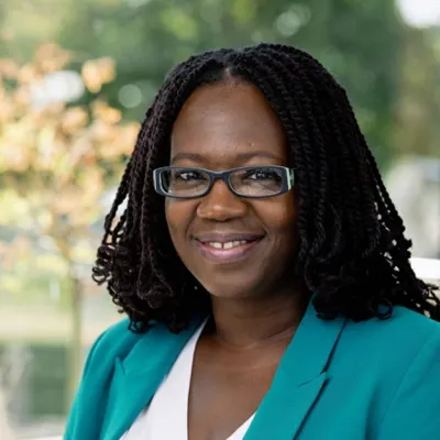 A person smiling with medium length black hair and glasses. They are wearing a teal jacket and white top, and are standing in front of trees.