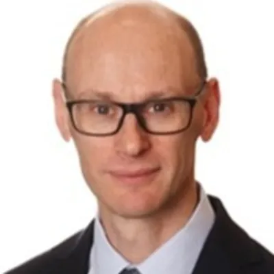 A person smiling in front of a white background. They are wearing glasses, a suit jacket and a white shirt and tie.