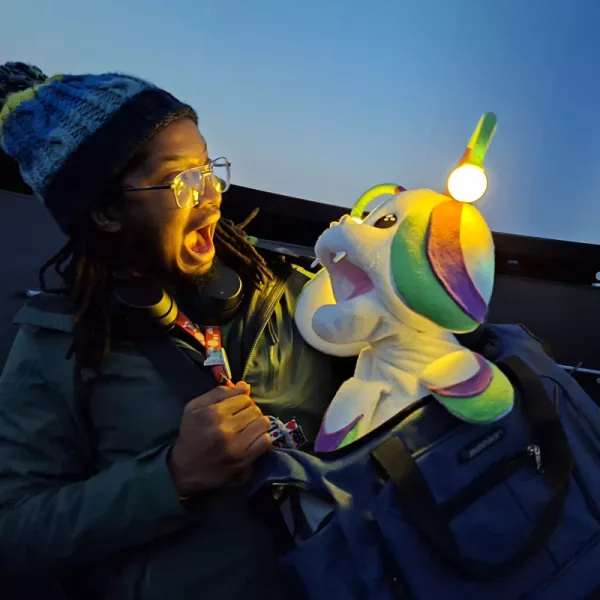 A person is stood in a dark room facing a puppet with their mouth wide open. The puppet is facing back at them with its mouth wide open. The puppet is white, green, and purple and resembles a mushroom-like creature. It has antenna which light up and little white fangs.