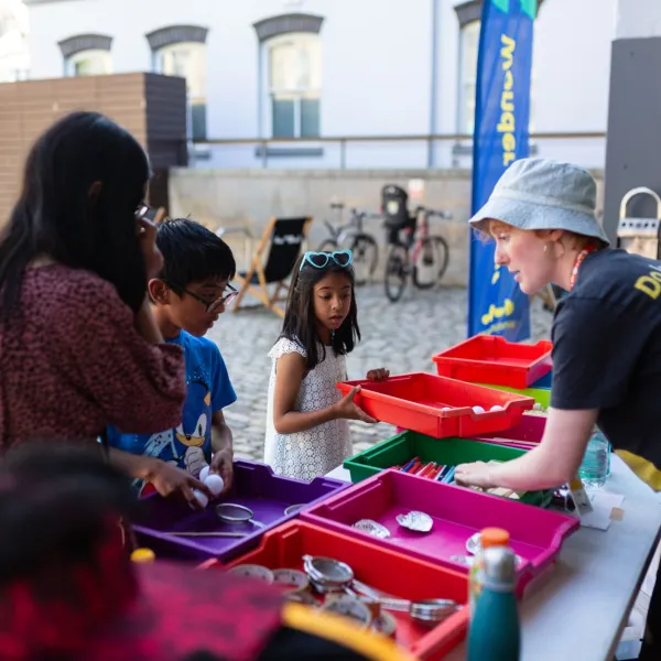 A family group of 3 with a We The Curious staff member, looking at things in a series of coloured trays in an outdoor space