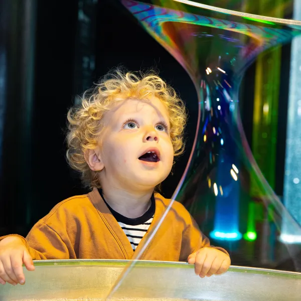 A toddler with blonde curly hair looking up at a giant bubble with mouth wide open