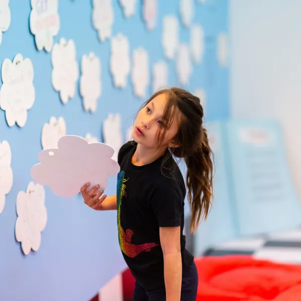 A child at an interactive learning exhibit, holding a cloud-shaped cutout next to a wall filled with similar cutouts.