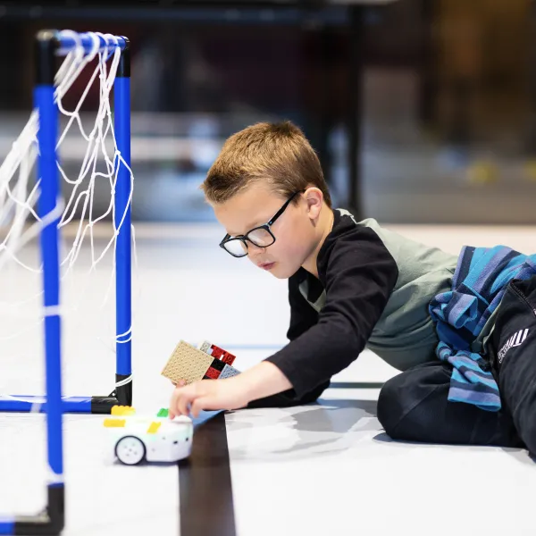 A child laying on the floor, pressing a small round wheeled robot, within a football goal