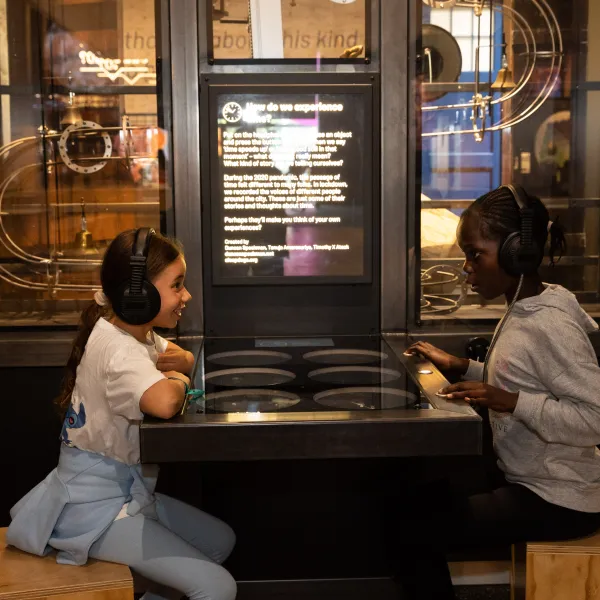 Two children are sat in a science museum opposite each other with a table in the middle of them. They are both wearing headphones 