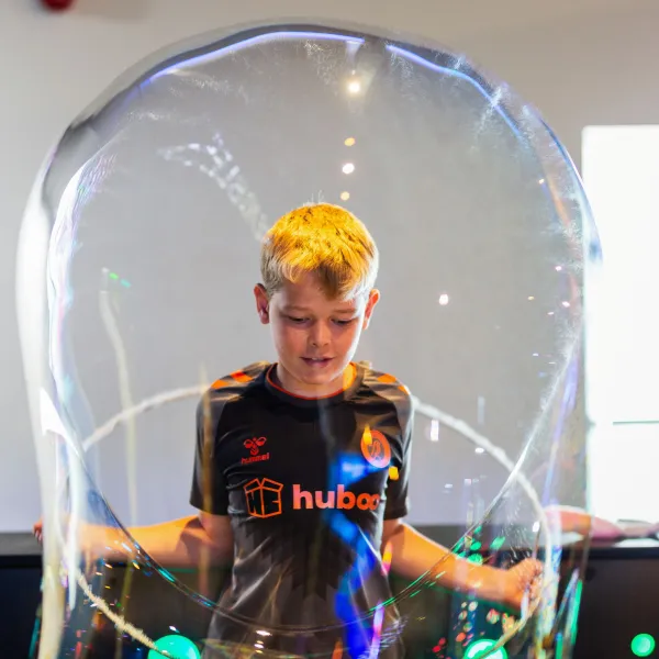 An older child is stood in a science centre, pulling a giant bubble over their head.