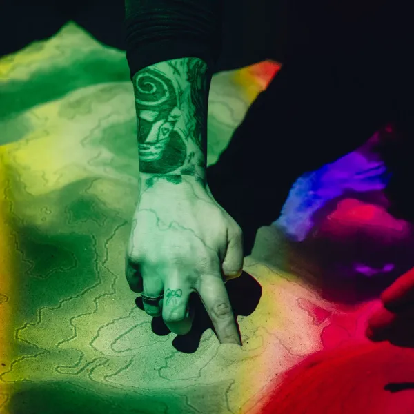 A person's tattooed arm reaching out into some sand, which is covered in contour lines and rainbow-coloured lights 