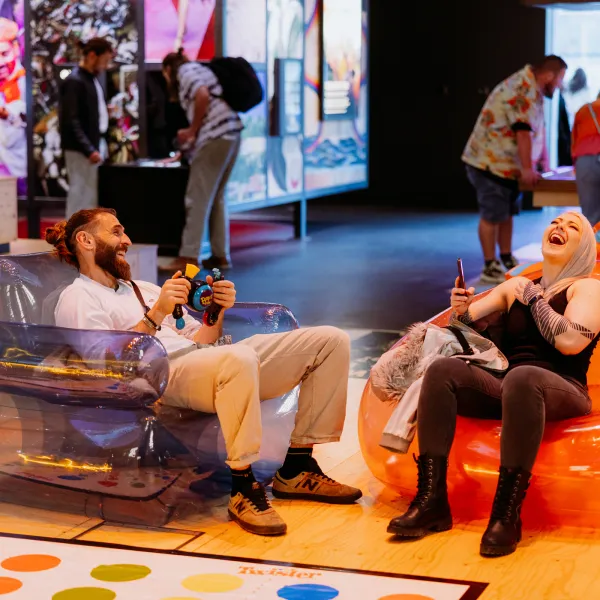 Two people are sat in the theatre of curiosity on bubble chairs, playing bopit and laughing.