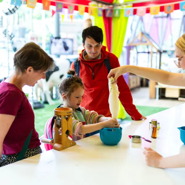 A child and two adults are stood behind a kitchen counter in a science museum. A member of staff is helping them squeeze oat milk from a cloth.