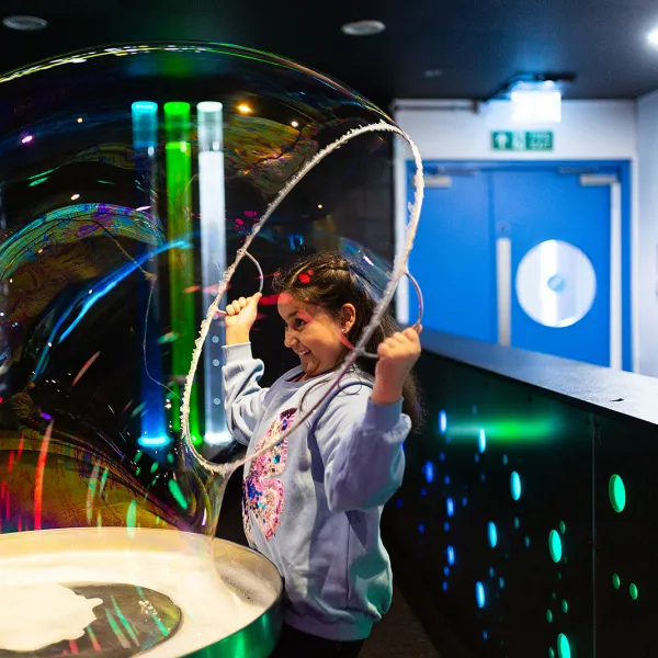 A child is in the middle of a science centre in a jumper pulling a sparkling bubble over their head.