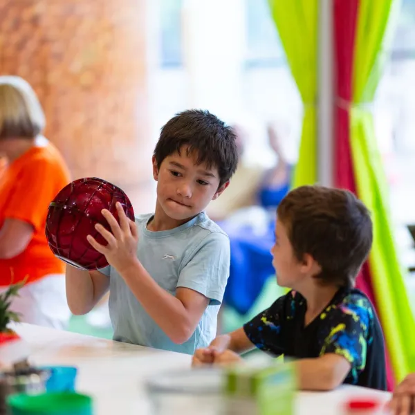 Two children are in the kitchen of a science centre. The older one is holding a big red ball and shaking it.