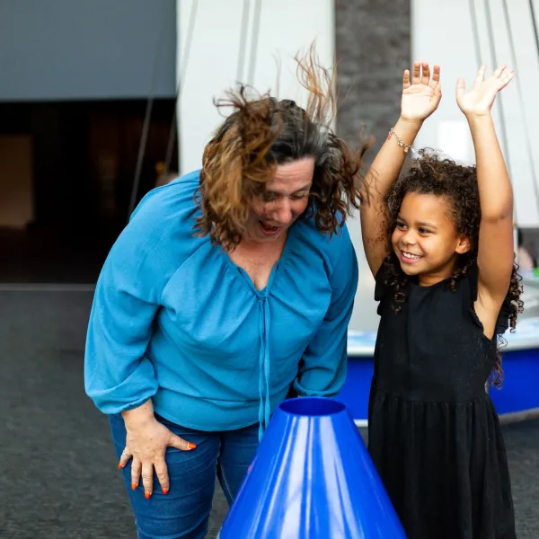 An adult and a child are stood in a science centre. The adult is putting their face over an air cannon and their hair is flying up. The child is stood next to them with their hands in the air. It's the Summer Holidays.