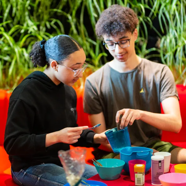 Two children are sat in the lab of a science centre on a red sofa. Looking down and playing with various kitchen equipment.