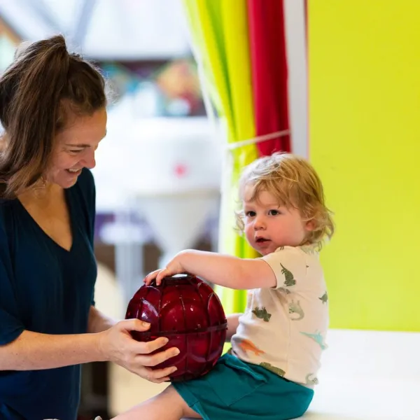 A child is seated on a counter top supported by an adult. They are holding a red sphere between them. 
