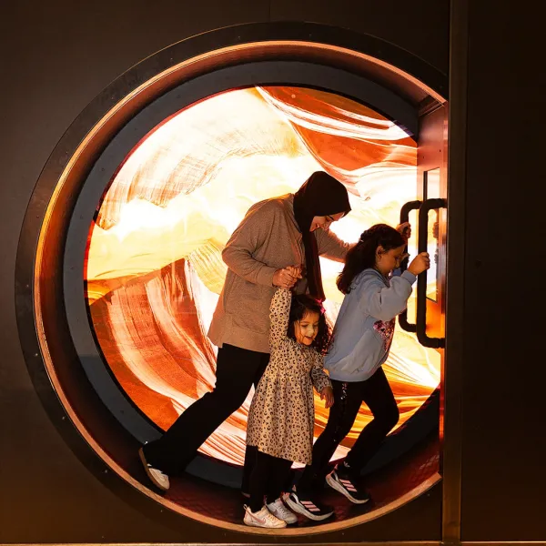 An adult and two children are pictured in the wheel in the sand exhibit at We The Curious. There is a colourful red and yellow background inside the wheel.