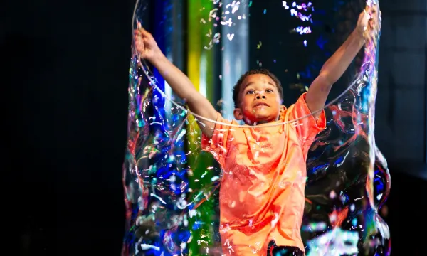 A child makes a large, colourful bubble using a large hoop which they are holding above their head.